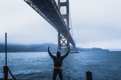 Rear view of man standing under bridge by bay against sky