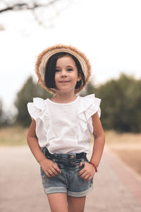 Smiling trendy kid girl 4-5 year old wear straw hat and summer white top with denim shorts outdoors