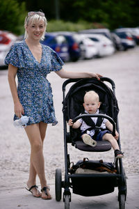 Mother in dress with 1 year old son in stroller in background of parking lot . summer
