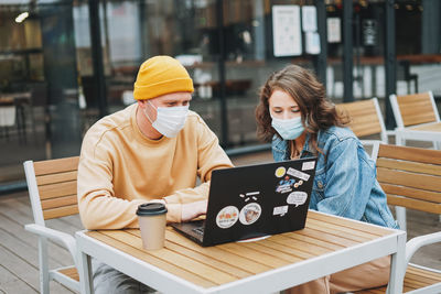 Stylish young couple freelancers in face mask working on laptop in street cafe, new normal