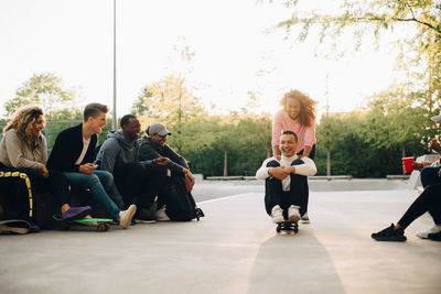 Friends looking at smiling woman pushing cheerful man sitting on skateboard at park