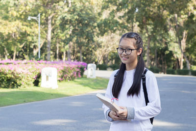 Portrait of smiling young woman holding book while standing on road