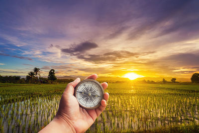 Person holding navigational compass on rice paddy against sky during sunset