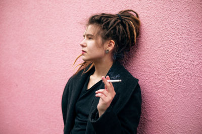 Hipster teen girl smoking in a warm black jacket leaning on pink loft brick wall