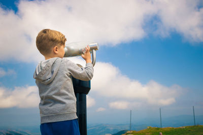 Boy looking through coin-operated binoculars against sky