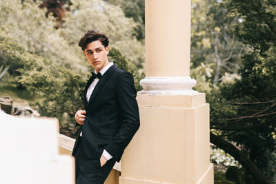 A beautiful young man, the groom in an elegant wedding suit, stands posing in the city's old park