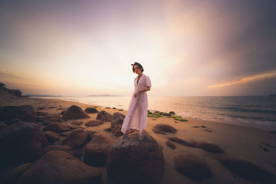 Woman standing on rock at beach against sky during sunset