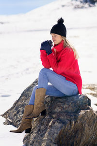 Full length of young woman sitting on rock on snow covered land