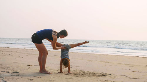 Full length of mother helping daughter in doing handstand on beach against clear sky