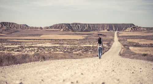 Rear view of woman walking on dirt road at desert against sky