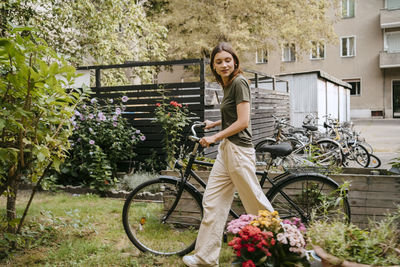Side view of young woman wheeling with bicycle while looking at flowers in garden
