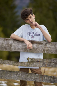 Portrait of young man standing by wooden fence on field