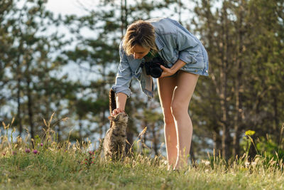 Woman photographer plays with lonely cat walking on forest meadow against blurry green trees