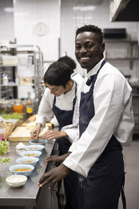 Portrait of smiling chef standing by female colleague garnishing food at commercial kitchen