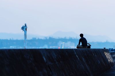 Rear view of man sitting on retaining wall against clear sky