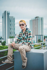 Portrait of young man wearing sunglasses and cap while sitting in city