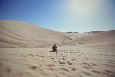 Rear view of man sitting on sand at desert against sky