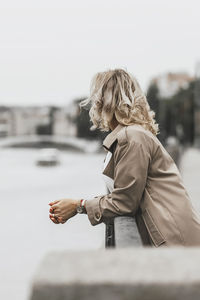 A middle-aged blonde woman with curly hair stands on the river embankment in the city.