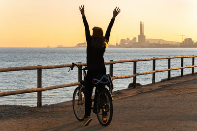 Rear view of woman on bicycle on sea against sky during sunset