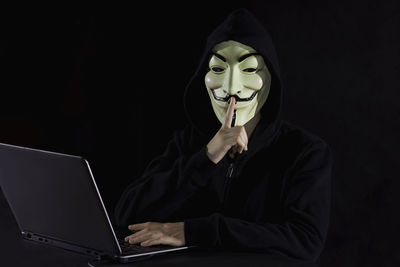 Portrait of computer hacker wearing mask with finger on lips against black background