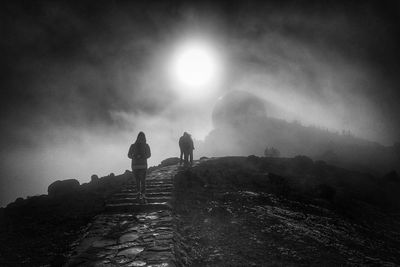 Low angle view of people walking on footpath against bright sun in sky