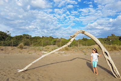 Cute girl standing by driftwood at beach against cloudy sky