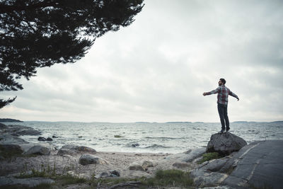 Rear view of wonderlust man standing with arms outstretched on rock by sea