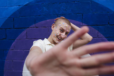 Happy non binary person with stickers on face showing stop gesture in front of colorful wall