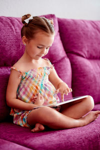 Cute girl using digital tablet sitting on pink sofa at home