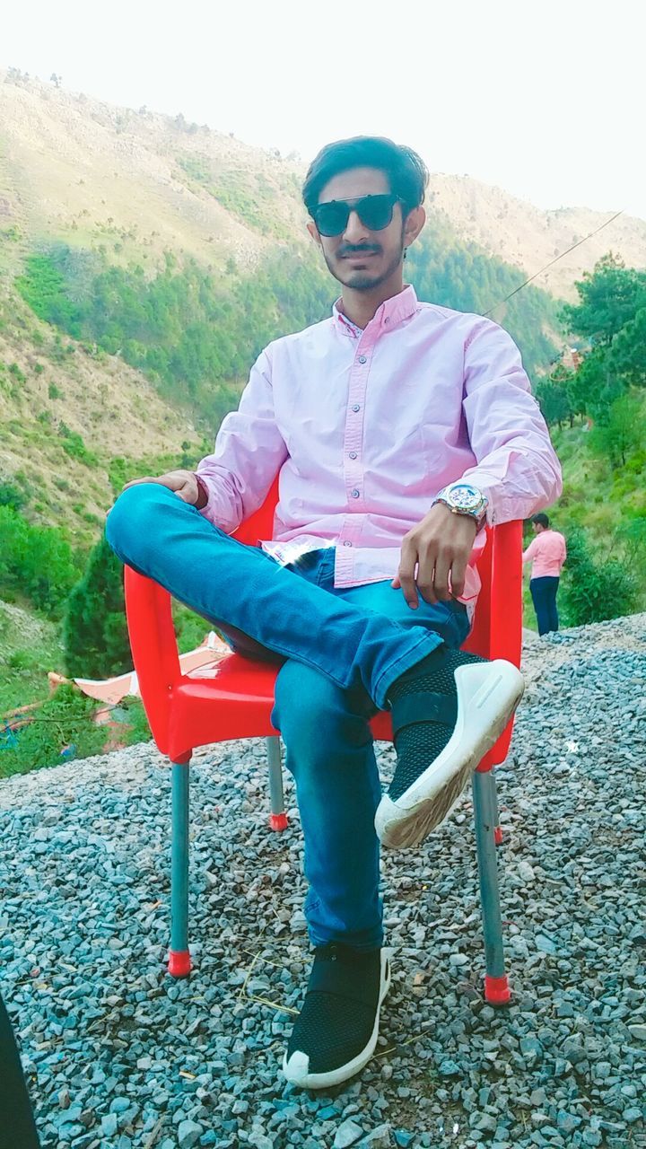 real people, one person, full length, young adult, leisure activity, lifestyles, young men, fashion, glasses, day, front view, sitting, sunglasses, nature, seat, casual clothing, mountain, men, clothing, outdoors