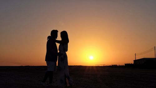 Side view of couple standing against orange sky during sunset