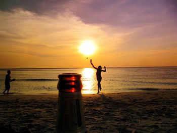 Beer bottle by playful silhouette people at beach against sky during sunset