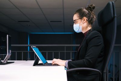 Side view of businesswoman specialist sitting at table and typing on keyboard on tablet with empty white screen while working at workplace