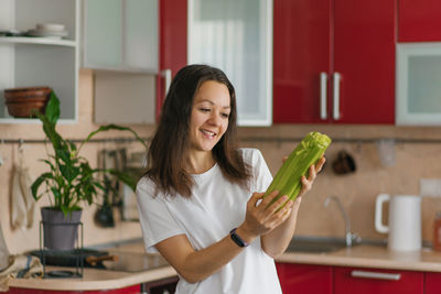 Smiling cheerful girl is standing in the kitchen with fresh vegetables and holding celery 