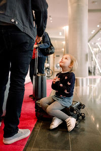 Full length of girl with hands clasped looking at father while sitting on suitcase in hotel