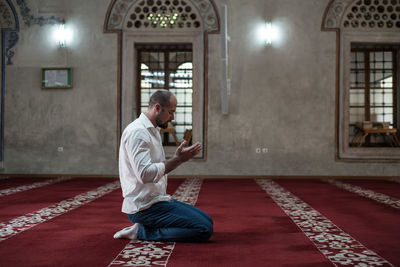 Side view full length of man praying while kneeling on carpet in mosque