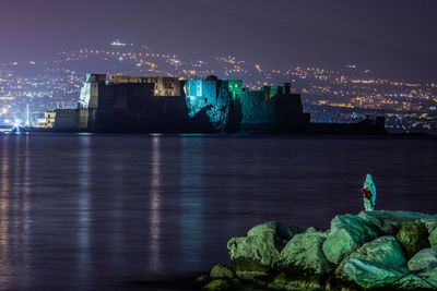 Illuminated buildings by sea against sky at night