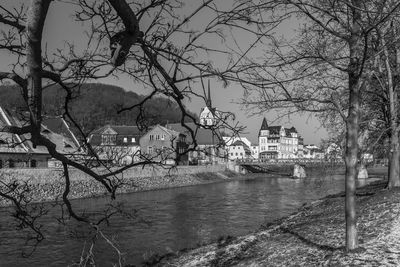 Bare trees by river and buildings against sky