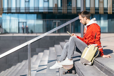 Stylish hipster in red jacket sitting on the steps. man surf in web with mobile phone. 