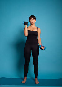 Full length of woman exercising with dumbbells against blue background