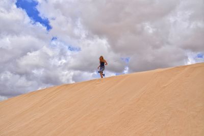 Low angle view of woman walking on sand dune against cloudy sky