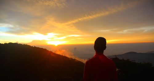 Rear view of man standing on mountain against cloudy sky during sunrise