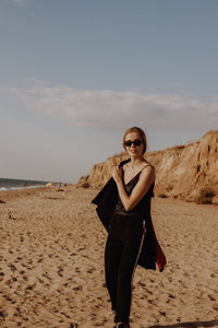 Portrait of young woman wearing sunglasses on beach against sky