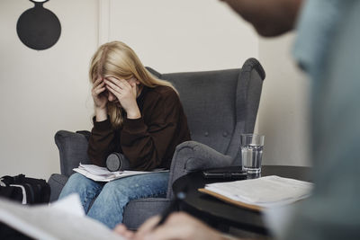 Blond teenage girl with head in hands sitting on chair while discussing with male counselor at school office