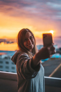 Smiling young woman taking selfie with mobile phone in balcony against sky during sunset