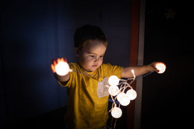 Curious little child playing with illuminated festive garland and standing in dark room in apartment