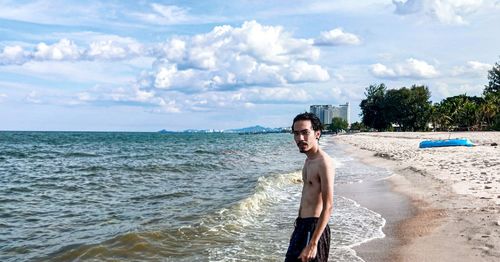 Portrait of shirtless man standing at beach against sky