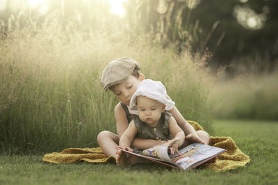 Little boy is reading from a book to his little sister sitting in the grass on a sunny summer day