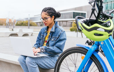 Colombian female freelancer sitting on concrete border near bicycle in modern city and doing online work