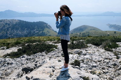 Full length of woman photographing while standing on rock against sky